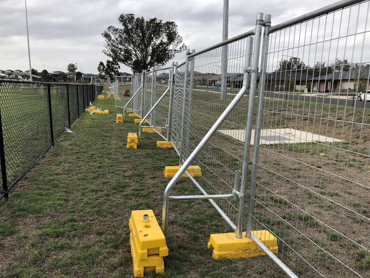 Temporary fencing with bracing bars and yellow stabiliser blocks installed on grassy site in suburban Australia